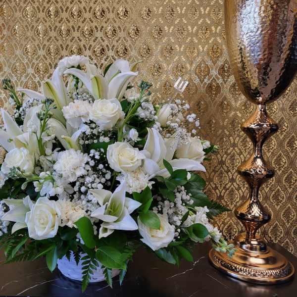 White lilies, roses, and carnations arranged in a white container with gold decor in the background.