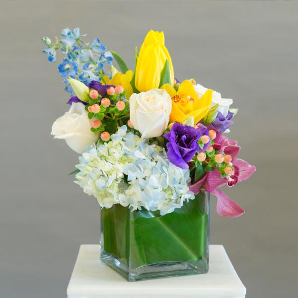 Mixed bouquet in a square glass vase with white, yellow, purple, and blue flowers