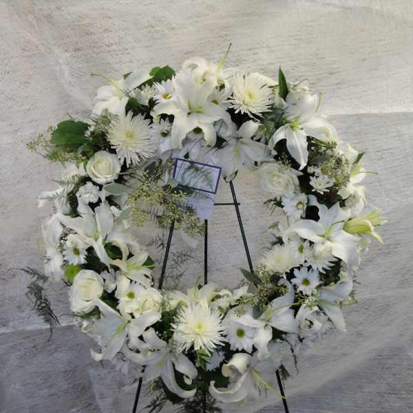 White floral wreath on a black stand with lilies and daisies