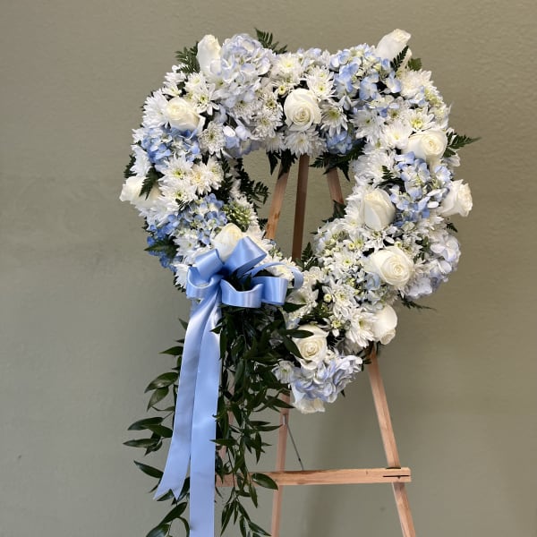 Heart-shaped floral wreath with white roses and blue flowers on a wooden stand