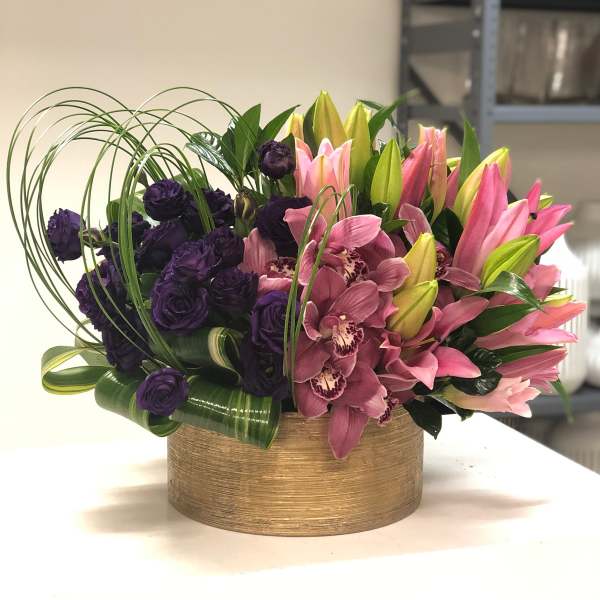 Low gold vase filled with pink lilies, purple roses, and pink orchids with looping decorative grass