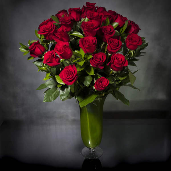 Tall arrangement of long-stem red roses in a green glass vase against a dark background