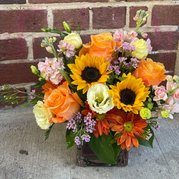 Mixed bouquet with sunflowers, orange roses, and pastel blooms in a glass vase