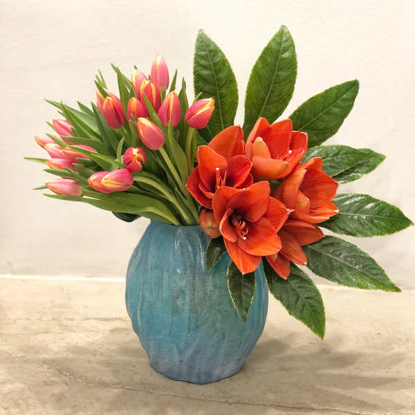 Asymmetrical vase arrangement with pink tulips, orange blooms, and a fan of glossy green leaves in a blue vase.