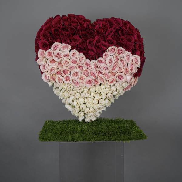 Heart-shaped rose arrangement in red, pink, and white on a grass-covered stand