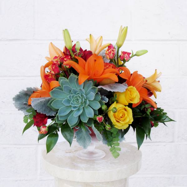 Bright bouquet of orange lilies, yellow roses, and a blue-green succulent in a glass vase.
