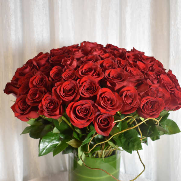 Large arrangement of red roses in a clear cylinder vase wrapped with green leaves.