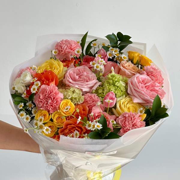 Handheld bouquet of pink, yellow, and orange flowers wrapped in clear paper