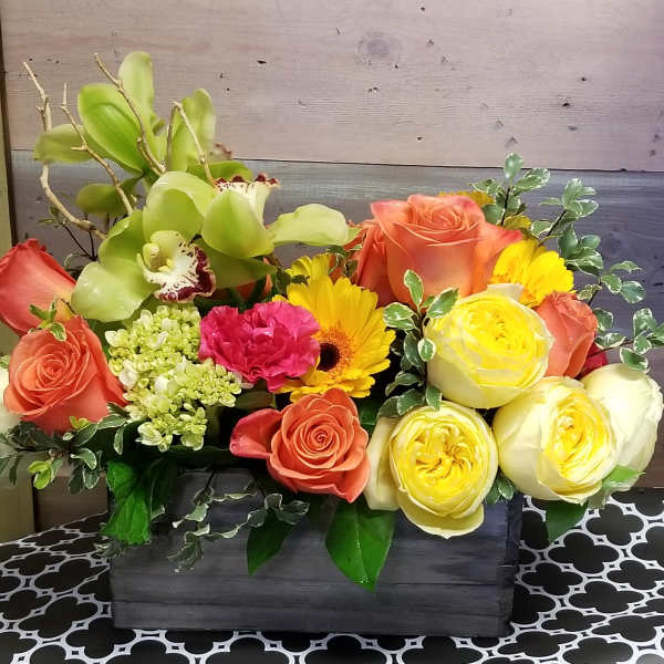 Low wood box arrangement with orange roses, yellow blooms, green orchids, and a pink carnation.