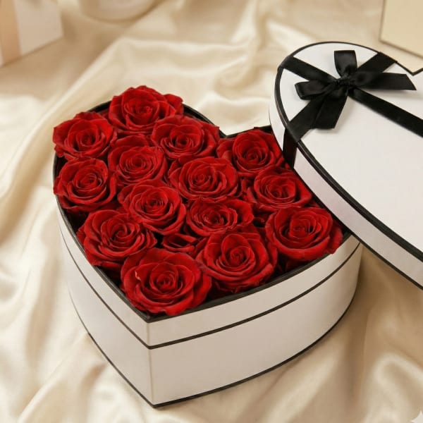 Heart-shaped white box filled with red roses, with a black-ribboned lid beside it.