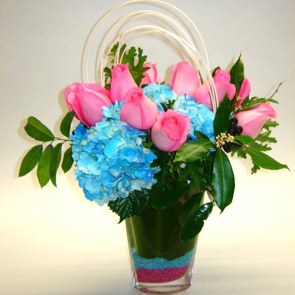 Pink roses and blue hydrangeas in a clear vase with arched handles