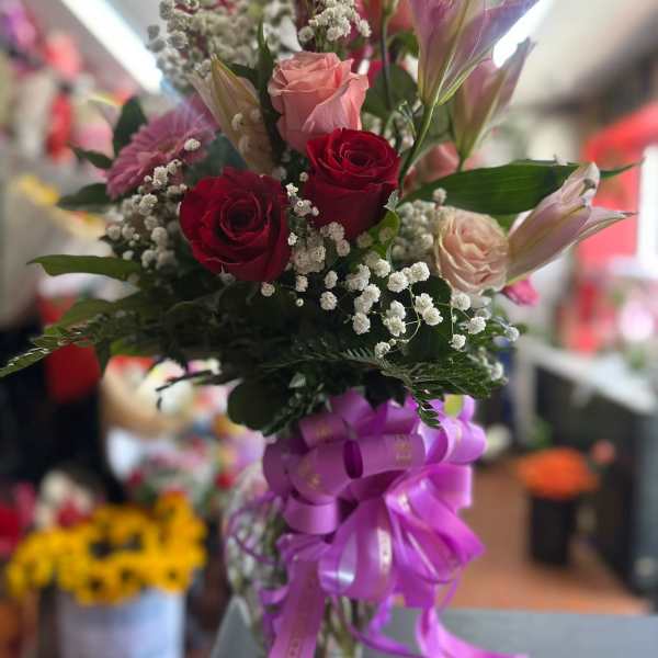 Tall vase arrangement of red and pink roses, pink lilies, and baby's breath with a large purple ribbon bow.