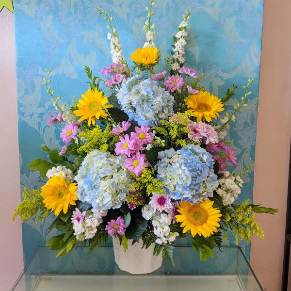 Large mixed flower arrangement in a white vase with blue hydrangeas and yellow daisies