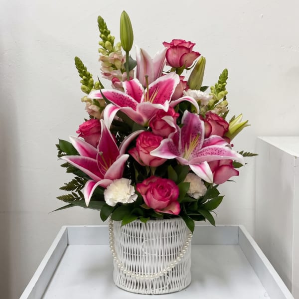 Pink lilies and roses arranged in a white basket vase with pearl trim