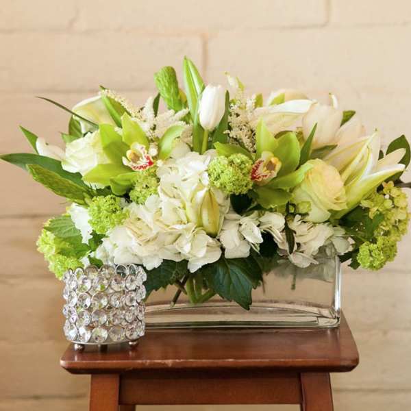 Low white and green floral arrangement in a clear glass vase beside a jeweled candle holder.