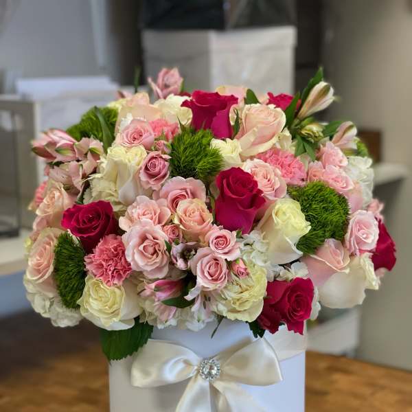 Round arrangement of pink and white roses with green accents in a white hatbox with satin bow