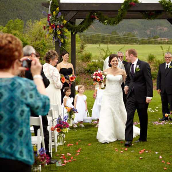 Bride and groom walking down an outdoor wedding aisle with floral arch and guests