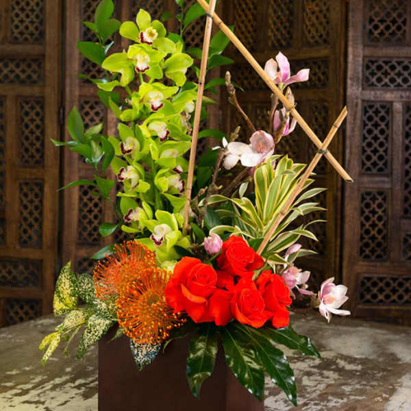 Tall floral arrangement with red roses, green orchids, and orange blooms in a square vase