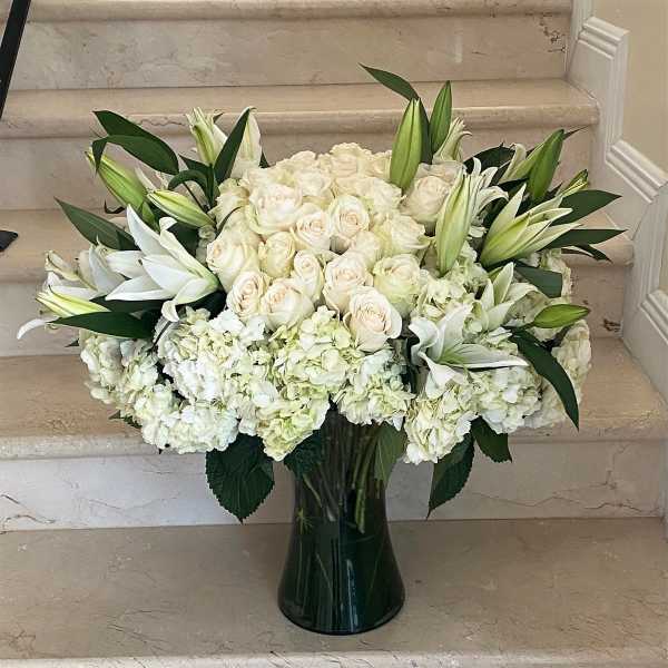 Large white arrangement of roses, lilies, and hydrangeas in a tall clear glass vase