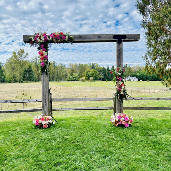 Floral arrangements decorate a wooden outdoor arch in a grassy field.