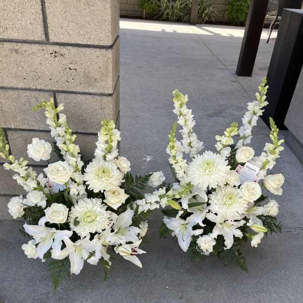 Two white floral arrangements with lilies, roses, and chrysanthemums on the ground.