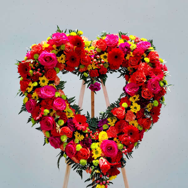 Heart-shaped floral wreath on a wooden stand with red, pink, and yellow flowers