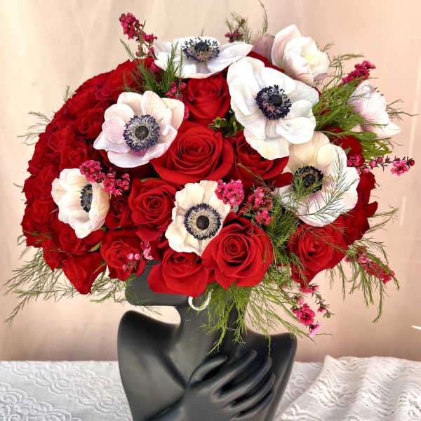 Red rose and white anemone arrangement in a black sculptural face vase on a lace-covered table.
