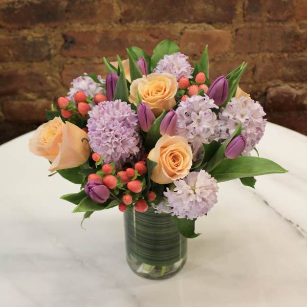 Bouquet of peach roses, lavender blooms, and purple tulips in a glass vase