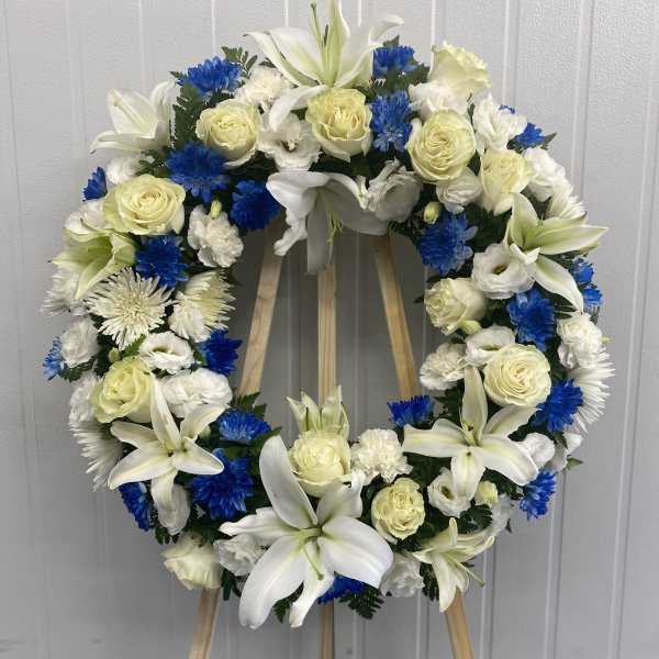 Circular funeral wreath with white lilies, cream roses, and blue flowers on a stand