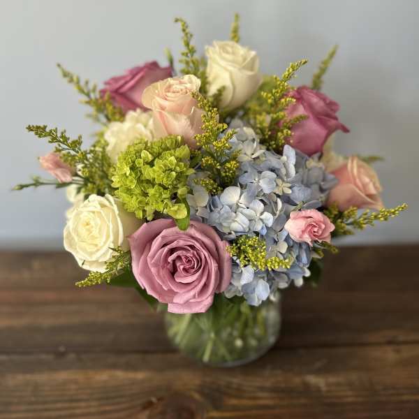 Round arrangement of pink and white roses with blue hydrangeas in a clear glass vase