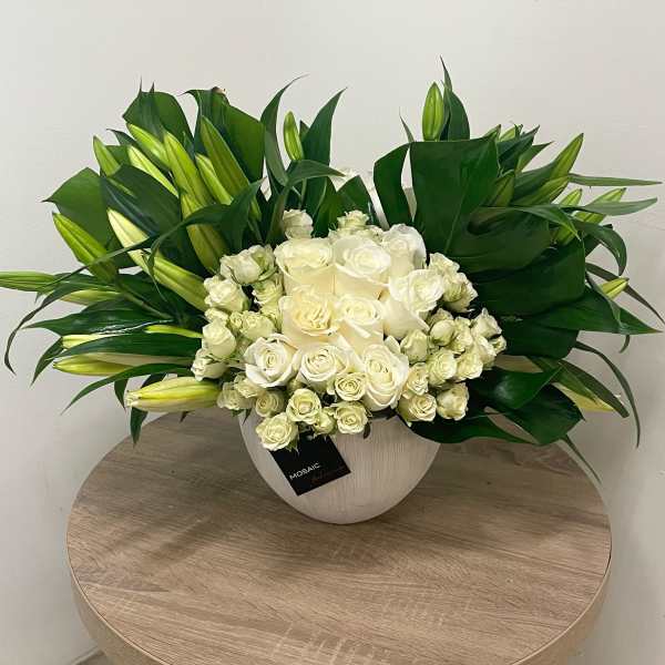 Low white bowl of white roses and white lily buds framed by large dark green leaves on a wooden table