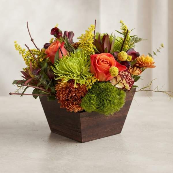 Mixed floral arrangement in a dark wooden container with orange roses and green blooms