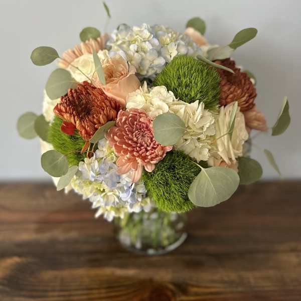Round arrangement of peach roses and mums with white and blue hydrangeas in a clear glass vase