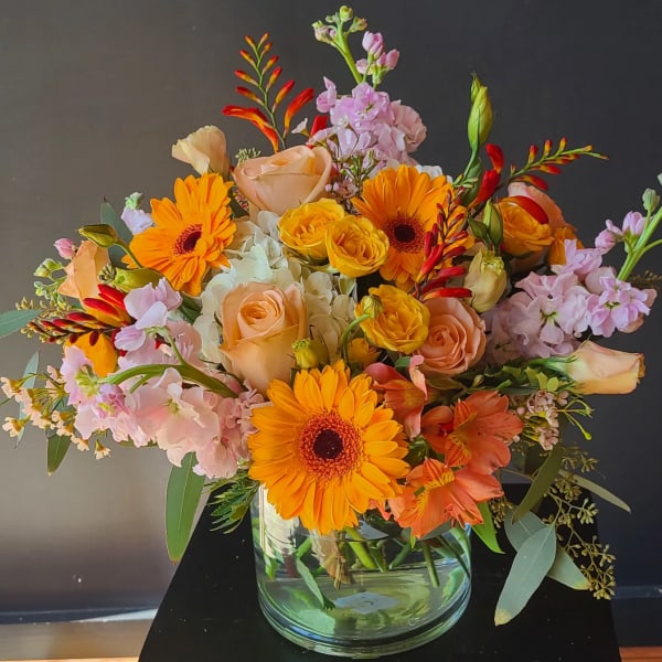 Mixed bouquet of orange gerbera daisies, peach roses, and pink blooms in a glass vase