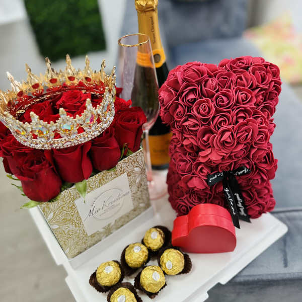 Red rose arrangement with a jeweled crown, champagne bottle, heart box, and chocolates