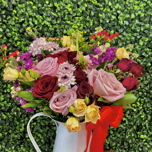 Mixed bouquet of roses and small blooms in a white metal pitcher with a red ribbon