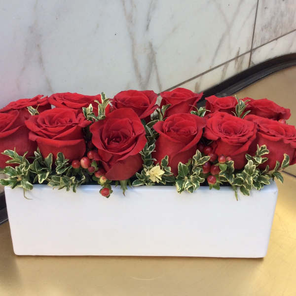 Red roses arranged in a white rectangular vase with small variegated greenery