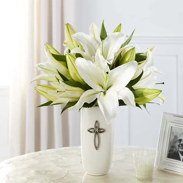 White lilies arranged in a tall white vase with a cross emblem