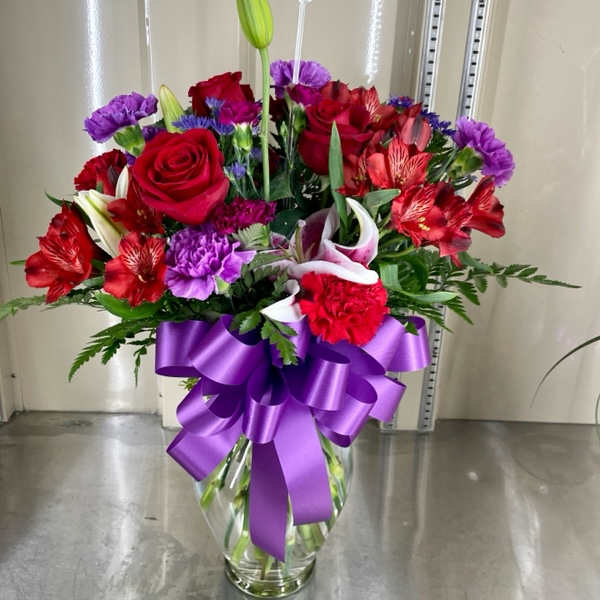 Bouquet of red roses, lilies, and purple flowers in a glass vase with a purple ribbon