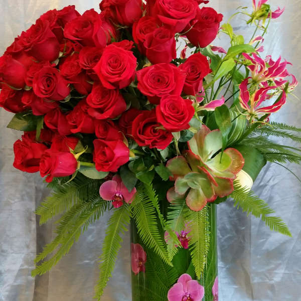 Red roses arranged in a tall glass vase with pink orchids and green foliage.