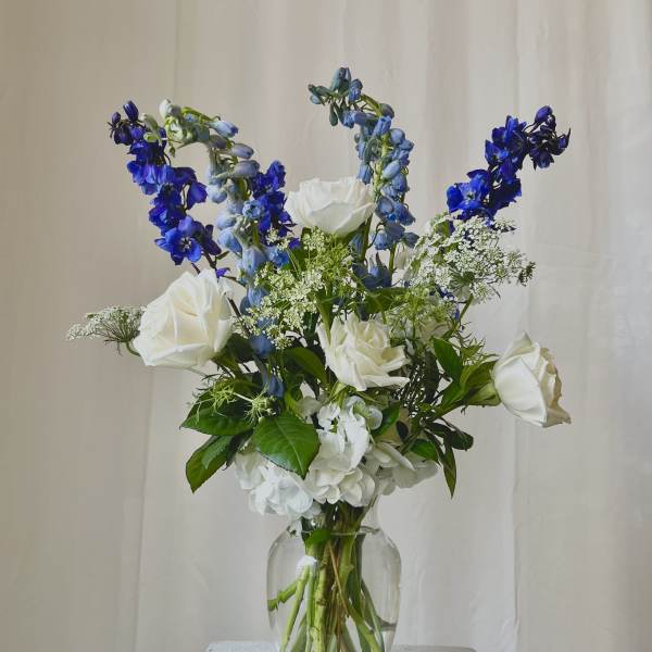Tall arrangement of white roses and hydrangea with blue delphinium in a clear glass vase