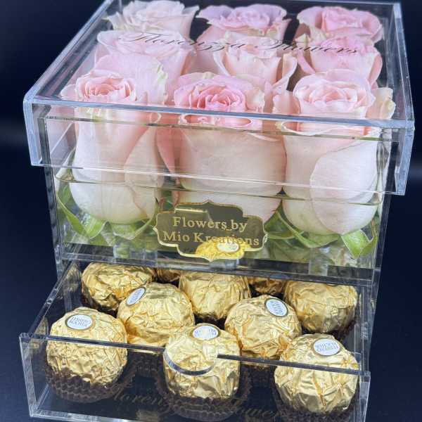 Pink roses in a clear box above gold-wrapped chocolates.