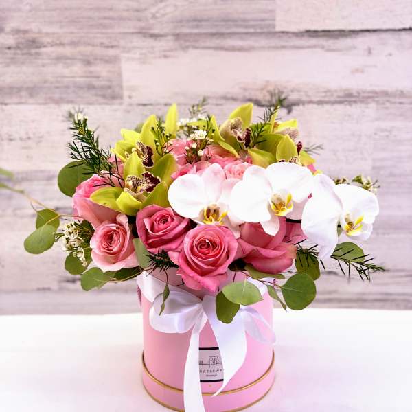 Pink roses and white orchids arranged in a pink hat box with a ribbon