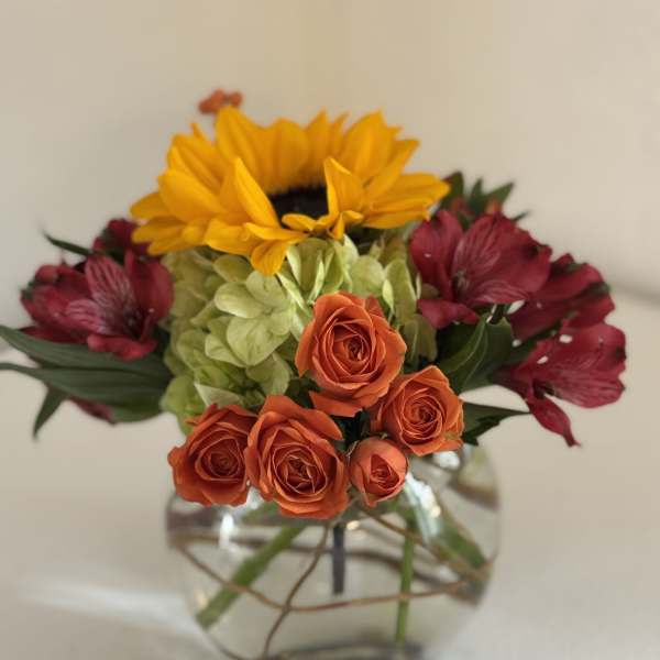 Mixed bouquet with orange roses, yellow sunflower, green hydrangea, and burgundy flowers in a glass vase