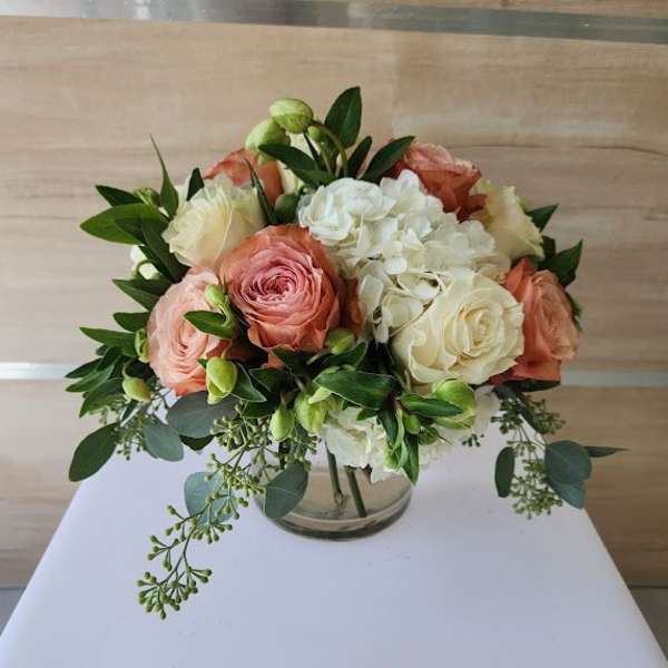 Short round bouquet of peach and cream roses with white hydrangeas in a clear glass vase