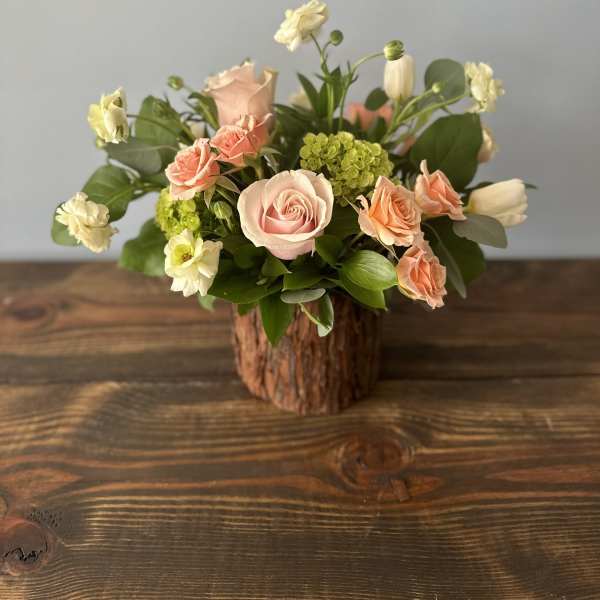 Short rustic arrangement of peach roses and white flowers in a bark-covered vase on a wooden table