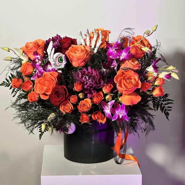 Orange and purple floral arrangement with roses, orchids, and Halloween skeleton hand accents in a black container