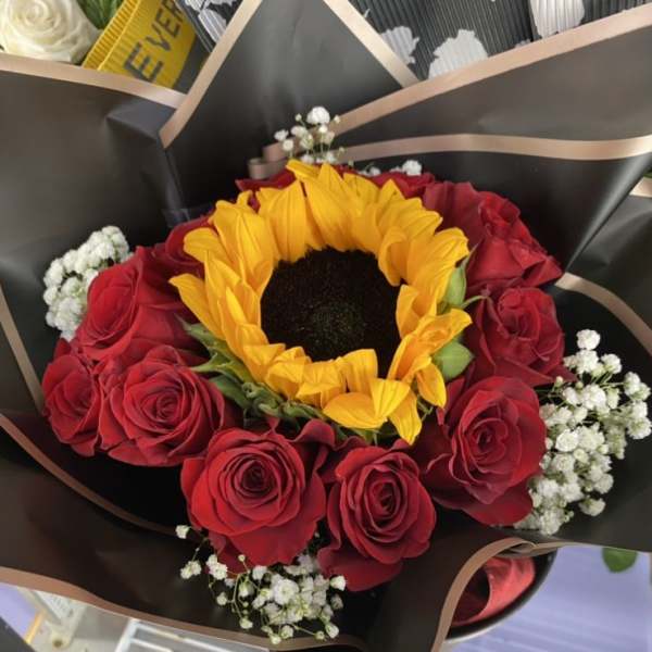 Hand-tied bouquet with a large yellow sunflower, red roses, and white filler flowers in black wrapping paper