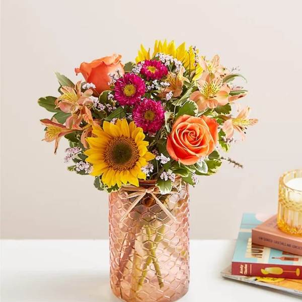 Bouquet of sunflowers, orange roses, and pink daisies in a textured glass vase