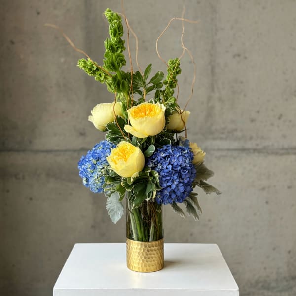 Yellow roses and blue hydrangeas in a gold vase with curly branches